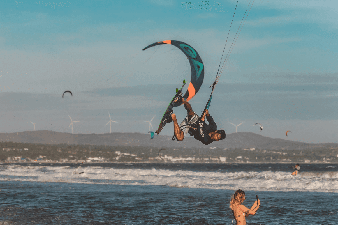 Canh, an IKO instructor at C2SKY, performing a high kite trick in the air on a windy day in Mui Ne, Vietnam, with kites and waves in the background.